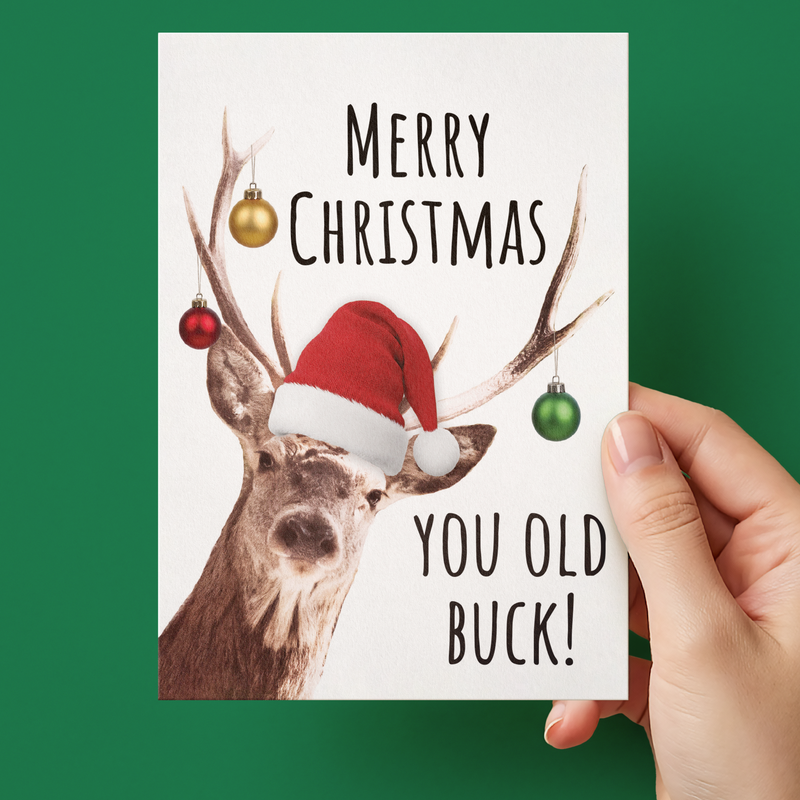A hand holding a white Christmas greeting card with a reindeer illustration wearing a Santa hat and the text 'MERRY CHRISTMAS YOU OLD BUCK!' against a solid green background.